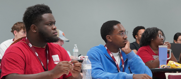 Two young men one with a red shirt and the other with a blue shirt listening to a discussion in a classroom setting.