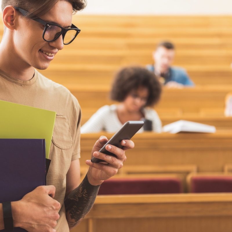 Outgoing young man is typing message on his smartphone while standing in auditorium. He is holding books and smiling. Copy space